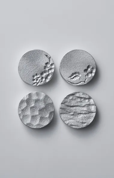Sand Mold Casting Four round, textured silver metal discs are arranged in a square on a plain, light gray background. Each disc has a unique surface pattern, including waves, dimples, and small raised or indented areas.