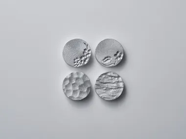 Four round, textured silver metal discs are arranged in a square on a plain, light gray background. Each disc has a unique surface pattern, including waves, dimples, and small raised or indented areas.