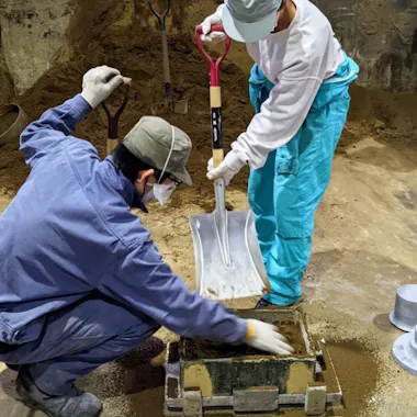 Sand Mold Casting Two people wearing masks, gloves, and work clothes fill a rectangular mold with sand using a large shovel in an industrial setting. Metal parts and tools are visible around them.