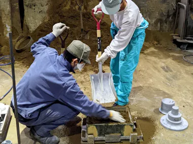Two people wearing masks, gloves, and work clothes fill a rectangular mold with sand using a large shovel in an industrial setting. Metal parts and tools are visible around them.