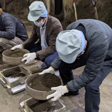 Sand Mold Casting Three people wearing blue caps, jackets, gloves, and face masks are crouched on the ground, sifting soil through metal sieves into trays inside an indoor, industrial setting. Shovels and piles of soil are visible nearby.