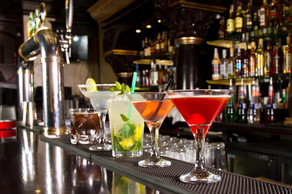 A row of colorful cocktails on a bar counter, including a martini, mojito, whiskey on the rocks, and a cosmopolitan. The bar background features shelves filled with various bottles of liquor and a large beer tap.
