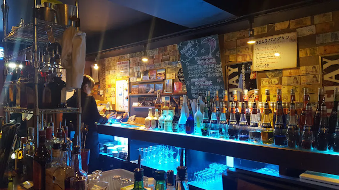 A dimly lit bar interior featuring a bartender standing behind a counter stocked with various bottles of liquor, illuminated by a blue light. Shelves of additional bottles and a mix of currency notes and menu boards cover the brick wall behind.