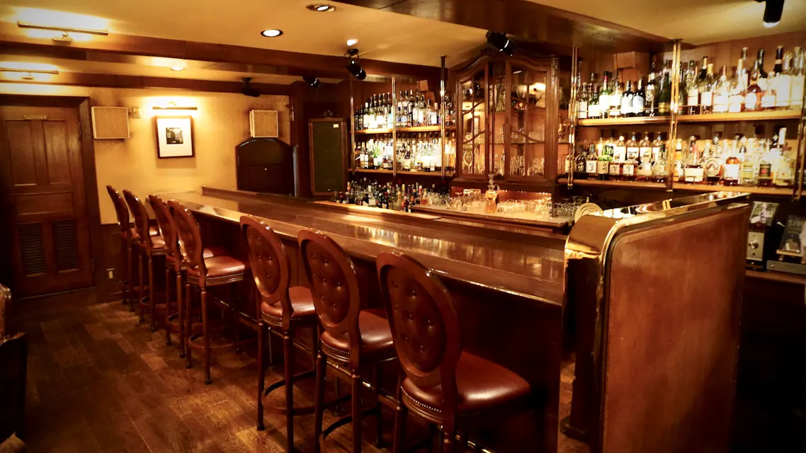 A cozy, dimly lit bar features a polished wooden counter with high-back leather chairs lined up against it. Shelves filled with assorted bottles of liquor adorn the back wall, creating a warm and inviting atmosphere. A framed picture hangs on the beige wall to the left.