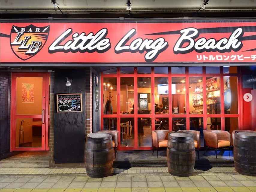 Street view of "Little Long Beach" bar with a red and black sign above the entrance. The facade features large windows and wooden barrels outside. The interior is warmly lit, showcasing a cozy seating area and a well-stocked bar. Japanese text is also on the sign.