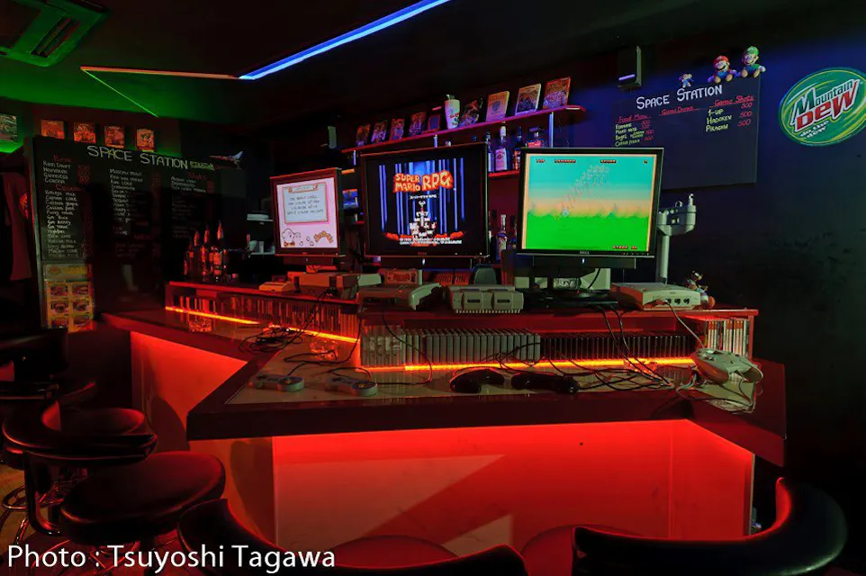A brightly lit, retro-themed bar features a couple of arcade machines on the counter. The dimly lit room is accentuated with neon red lighting. Shelves behind the bar display various gaming memorabilia and drinks. The overall ambiance is lively and nostalgic.