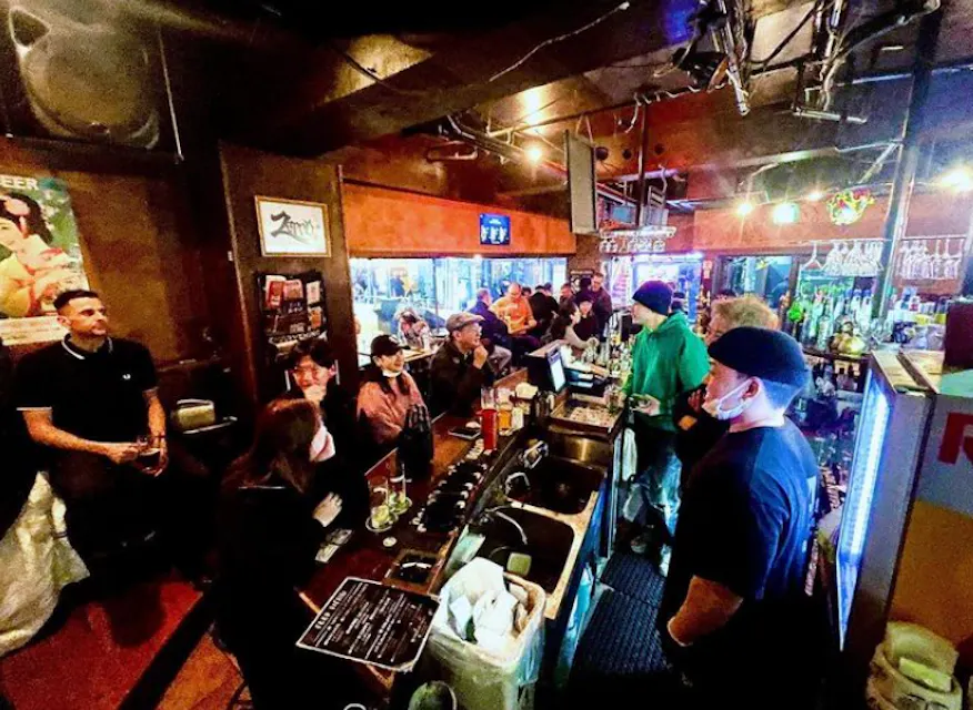 A vibrant bar interior filled with people enjoying drinks and conversation. Patrons are seated at the bar and at tables, while several staff members serve behind the bar. The atmosphere is lively with colorful lighting and decor, including a neon sign in the background.