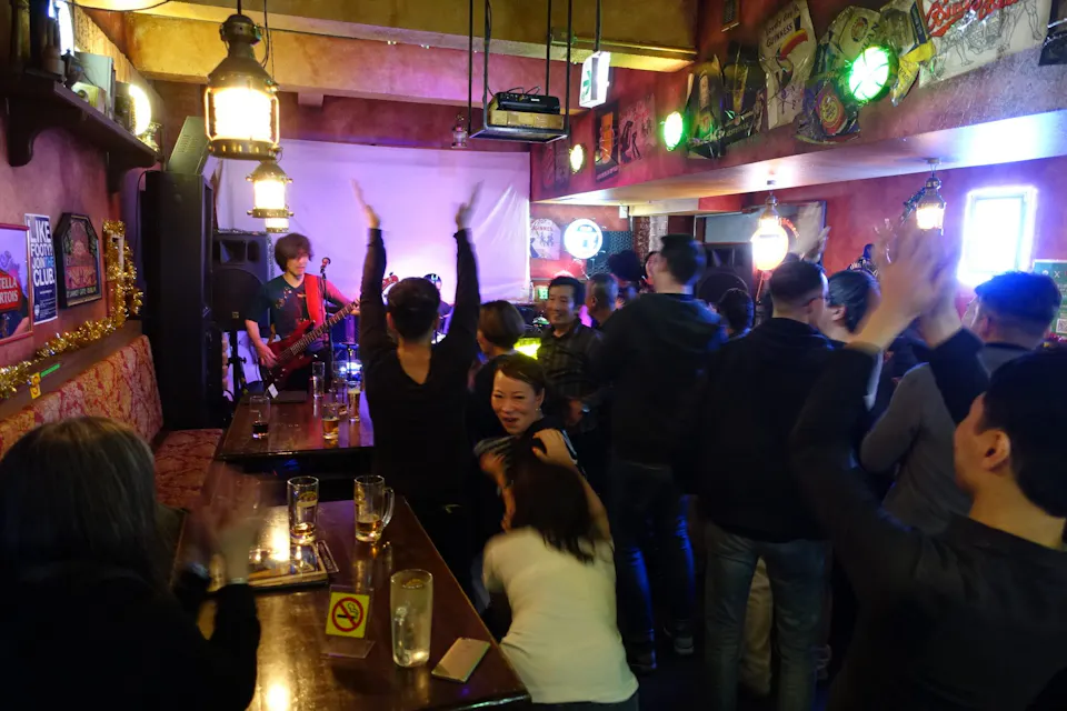 A live band performs on stage in a cozy, dimly lit bar, with a guitarist in a red shirt at the center. The audience cheers and raises their hands in excitement. Tables with drinks occupy the foreground, and various decorative items adorn the walls.