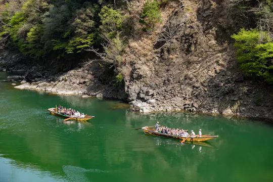 Two wooden boats with tourists glide on a calm green river beside rocky, tree-covered cliffs under daylight. The passengers appear to be enjoying a scenic nature tour.