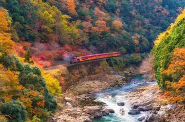 A vibrant red and yellow train travels through a forest with colorful autumn foliage, crossing a river gorge with rocky banks, while people in boats paddle along the river below.