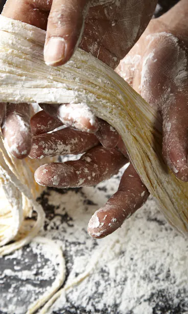 Handmade Noodles Hands coated in flour are stretching and shaping fresh, uncooked pasta noodles on a floured surface, with loose strands and scattered flour visible in the background.