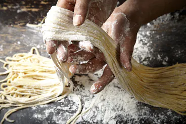 Hands coated in flour are stretching and shaping fresh, uncooked pasta noodles on a floured surface, with loose strands and scattered flour visible in the background.