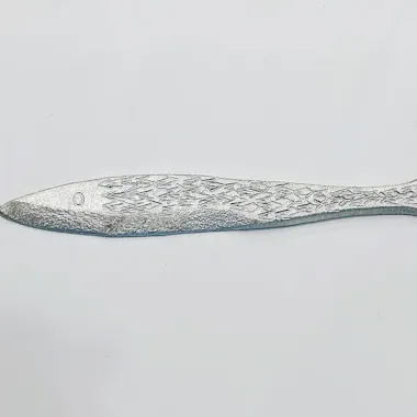 Sculpting Workshop A silver metal object shaped like a fish with engraved scale patterns and a small dot for an eye, placed on a plain white background.