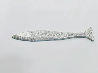 A silver metal object shaped like a fish with engraved scale patterns and a small dot for an eye, placed on a plain white background.