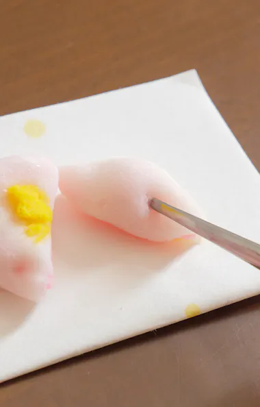 Confectionery Cutter A hand holding a piece of pink mochi with yellow filling on a white napkin, while another hand cuts it with metal chopsticks on a wooden table.