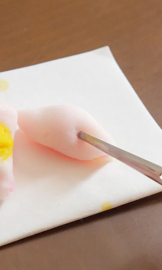 Confectionery Cutter A hand holding a piece of pink mochi with yellow filling on a white napkin, while another hand cuts it with metal chopsticks on a wooden table.