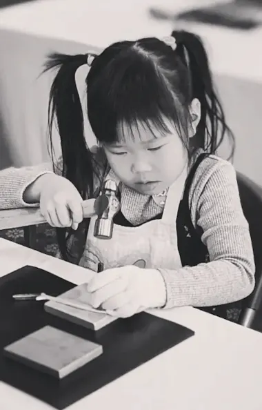 Engraving Workshop A young girl with pigtails focuses intently as she uses a hammer and metal tool to work on a craft project at a table. She wears a light-colored apron and sits among various crafting materials.