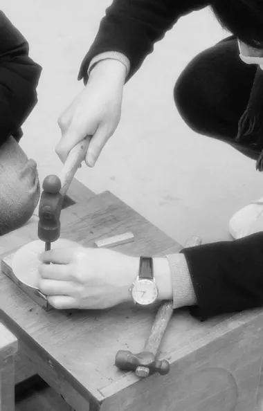 Engraving Workshop Two people sit close together. One holds a metal object steady on a small block while the other watches. The person with a watch uses a hammer and tool to work on the object. Both wear masks.