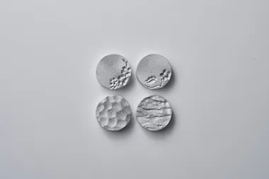 Four round silver objects with textured surfaces, each displaying a different raised pattern, are arranged in a square on a plain light gray background.