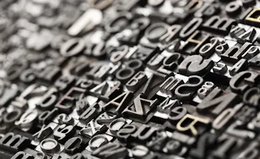 A close-up view of assorted metal letterpress type blocks arranged tightly together, featuring various letters, numbers, and symbols in different fonts and sizes, with a shallow depth of field.