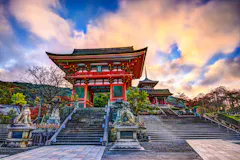 A vibrant red traditional Japanese temple gate with stone lion statues stands at the top of wide steps, framed by trees and a dramatic, colorful sky.