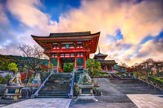 A vibrant red traditional Japanese temple gate with stone lion statues stands at the top of wide steps, framed by trees and a dramatic, colorful sky.