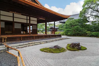 A traditional Japanese wooden building overlooks a peaceful Zen garden with raked gravel, moss, and rocks, surrounded by green trees under a partly cloudy sky.