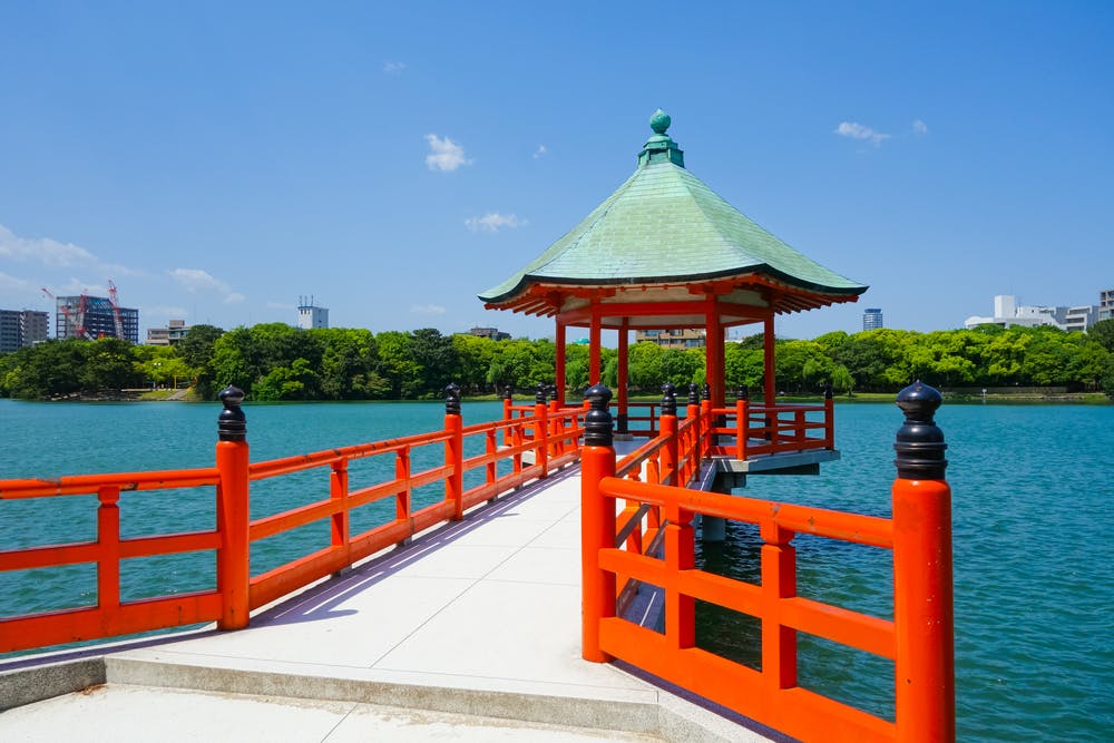 A traditional Japanese pavilion with a green roof sits over a calm blue lake, connected by a bright red wooden walkway. Trees and city buildings are visible in the background under a clear sky.