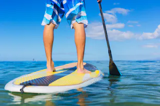 A person wearing blue and white board shorts stands barefoot on a paddleboard in calm, clear water, holding a paddle, with a bright blue sky and scattered clouds in the background.