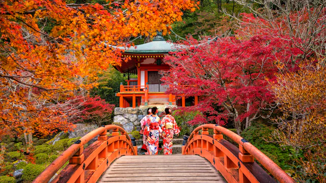 Two people in traditional kimonos walk across a red wooden bridge surrounded by vibrant autumn foliage, leading to a small Japanese pavilion in a lush garden.