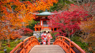 Two people in traditional kimonos walk across a red wooden bridge surrounded by vibrant autumn foliage, leading to a small Japanese pavilion in a lush garden.