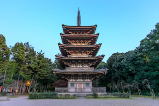 A traditional five-story pagoda stands surrounded by trees, with intricate wooden architecture and a tall spire, against a clear blue sky.