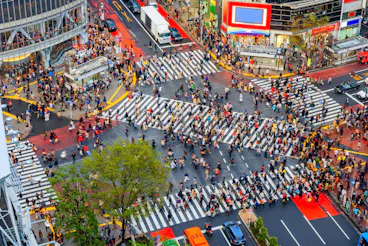 Aerial view of a busy urban intersection with hundreds of people crossing wide striped crosswalks in different directions. Bright buildings, vehicles, and a tree are visible around the crowded crossing.