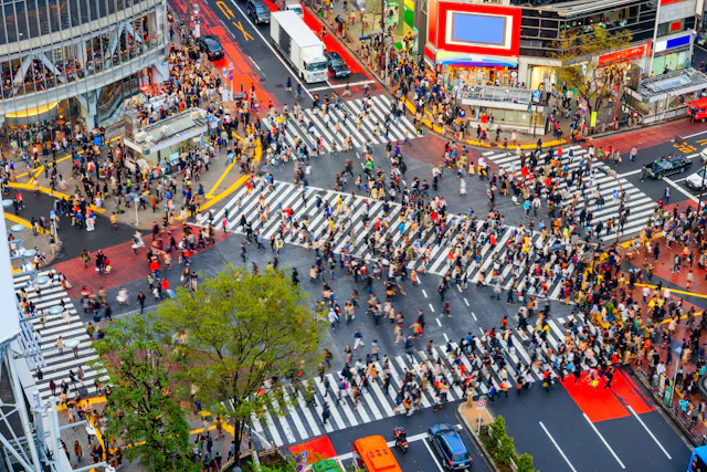 Aerial view of a busy urban intersection with hundreds of people crossing wide striped crosswalks in different directions. Bright buildings, vehicles, and a tree are visible around the crowded crossing.