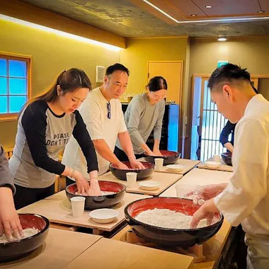 Soba Making A group of people stands around tables mixing dough in large bowls, guided by a chef in white, participating in a cooking class in a warmly lit room.