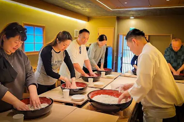 A group of people stands around tables mixing dough in large bowls, guided by a chef in white, participating in a cooking class in a warmly lit room.