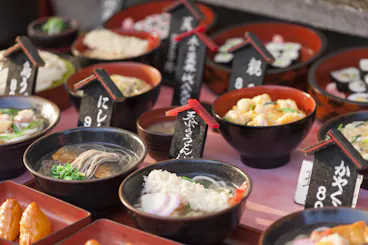 A display of various Japanese noodle and rice dishes in bowls with small black signs showing names and prices, likely outside a restaurant as sample food models to attract customers.