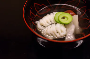 A lacquered bowl with clear broth, containing white fish, green cucumber slices, a piece of bamboo shoot, and a red plum, presented on a dark surface.