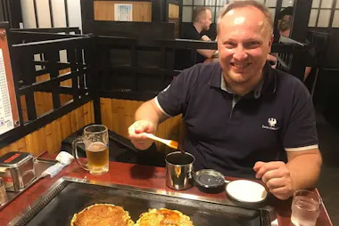 A smiling man sits at a restaurant table with a glass of beer, enjoying Japanese food. He holds a utensil and prepares food on a hot grill in front of him. The setting has wooden paneling and partitioned booths.