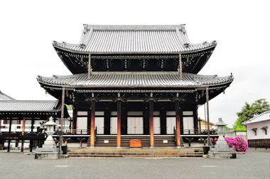 A traditional Japanese temple with a large, tiered roof, wooden pillars, and stone lanterns in front, surrounded by gravel and blooming bushes. The sky is overcast.