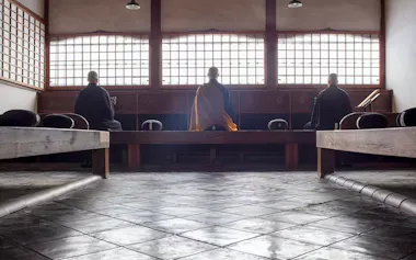Three people in robes sit meditating on cushions in a serene, minimalist room with large wooden windows and soft natural light. The floor is tiled, and benches line both sides of the space.