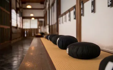 A row of black meditation cushions sits on a tatami mat bench in a quiet, traditional Japanese room with wooden walls and soft natural light.
