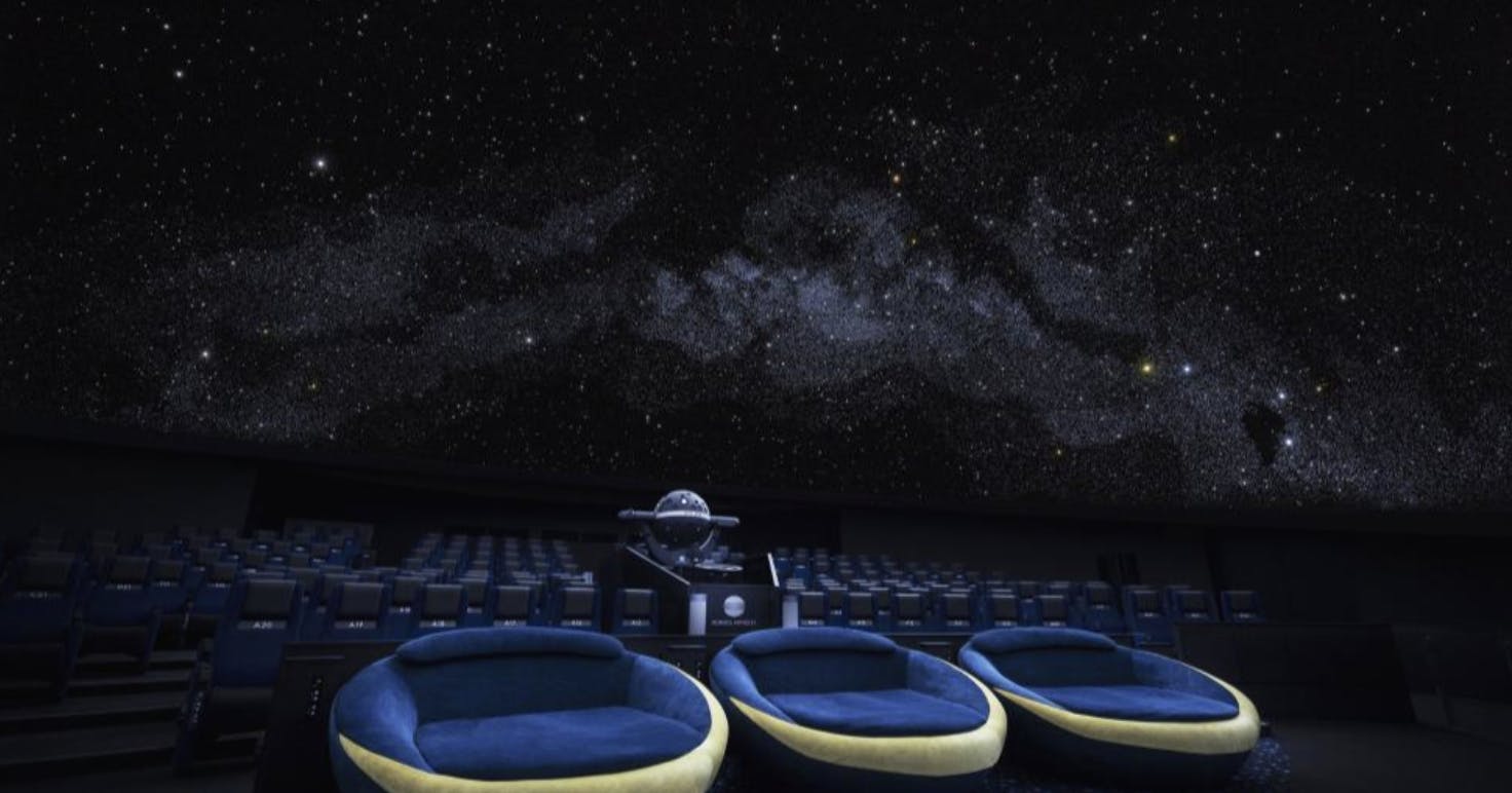 A planetarium interior with a star-filled projection on the domed ceiling, rows of seats, and three circular cushioned seats in the foreground facing the center of the room.