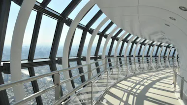 Panoramic view from inside a tall observation deck with curved glass windows, white ceiling, metal railings, and cityscape visible through the windows on a bright, sunny day.
