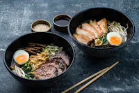 Two black bowls of ramen placed on a dark textured surface. One bowl contains ramen with sliced pork, soft-boiled egg, sprouts, and greens. The other bowl has ramen with sliced meat, soft-boiled egg, mushrooms, and greens. Chopsticks rest beside the bowls.