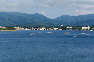 A serene blue lake with small floating platforms, set against a backdrop of green, forested mountains and a small town along the shoreline under a partly cloudy sky.