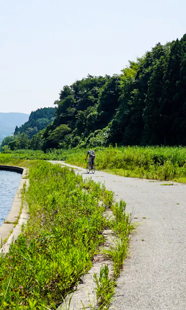 Sado Island A person rides a bicycle on a path beside a calm lake, bordered by green grass and trees, with mountains and houses visible in the background under a clear sky.