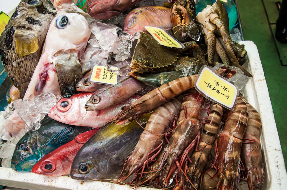 A selection of fresh seafood on display at a market stall, including fish, shrimp, and crabs. The seafood is laid on a bed of ice, with handwritten price tags in Asian script.