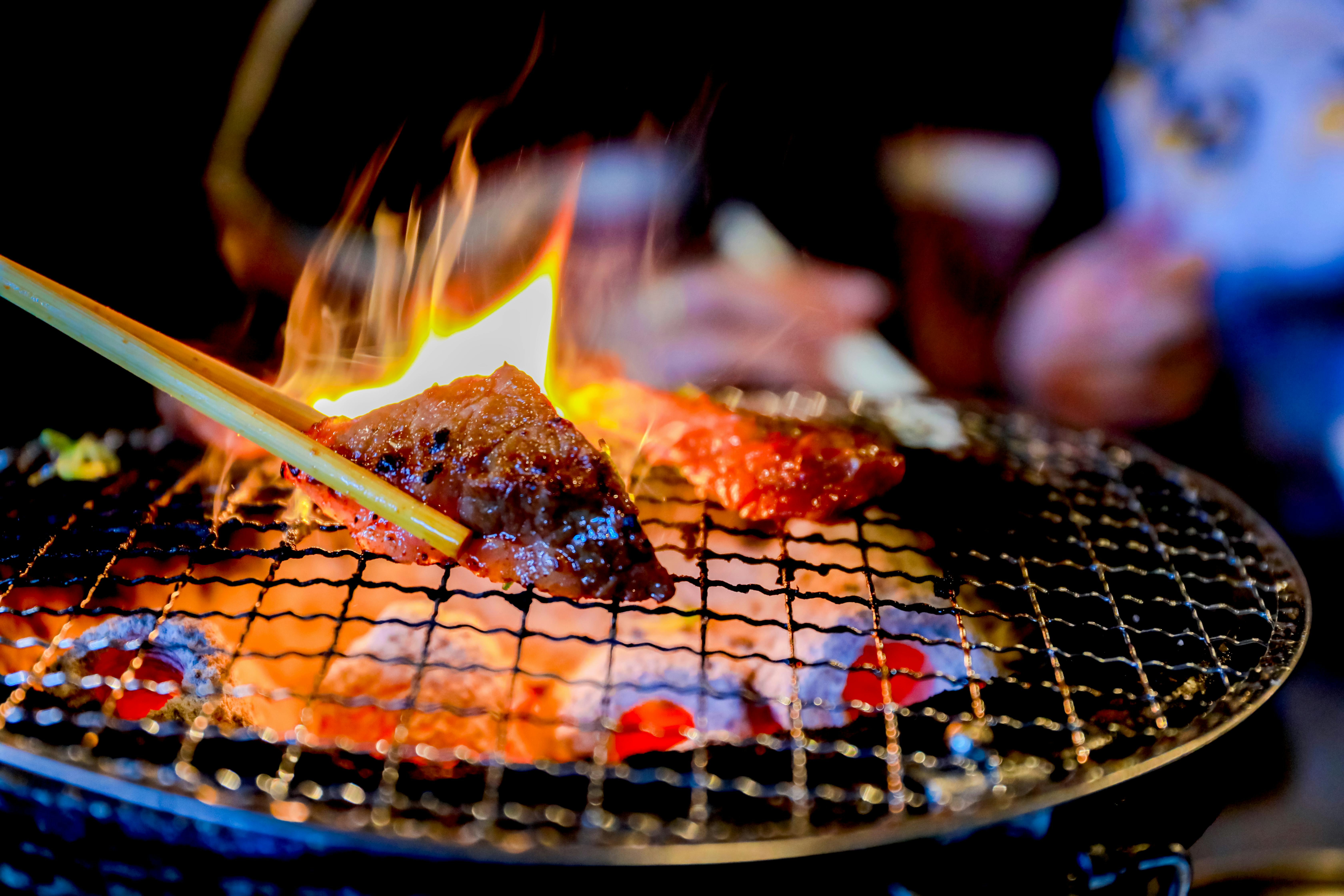 A close-up of meat being grilled over an open flame on a wire mesh grill, with chopsticks holding a piece of cooked meat. The fire is visible beneath the grill, and the background is blurred.