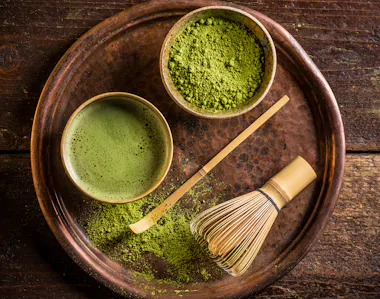 A wooden tray holds a bowl of matcha tea, a bowl of matcha powder, a bamboo whisk, and a bamboo scoop, all arranged on a rustic wooden surface.
